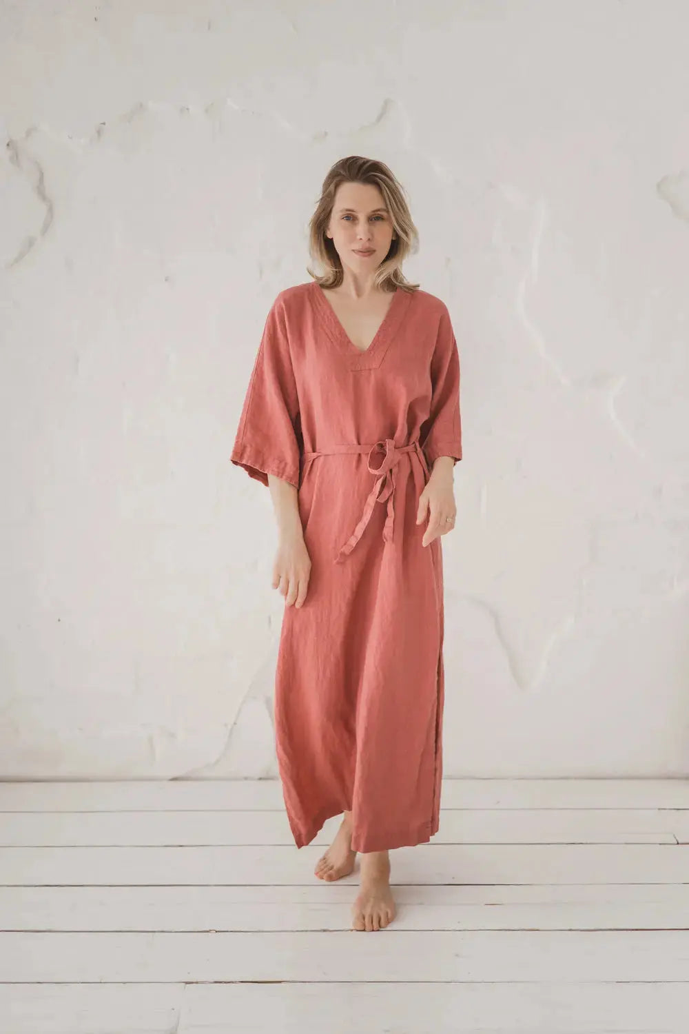 A woman wearing a dusty rose linen kaftan dress with a tie waist belt, standing barefoot on a white wooden floor against a textured white wall.