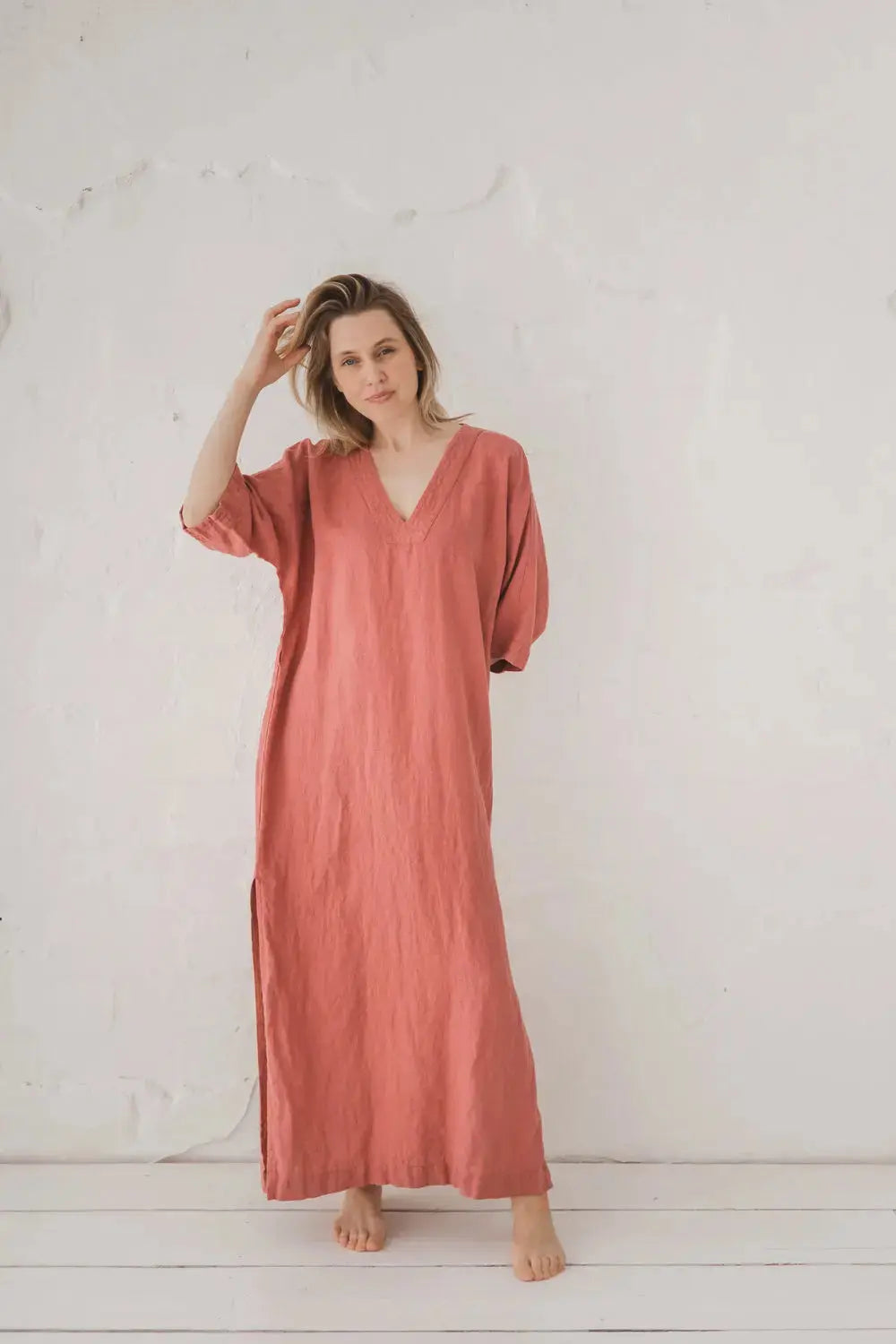 A woman wearing a loose, dusty rose pink kaftan dress with a V-neckline and elbow-length sleeves, standing barefoot against a textured white wall.