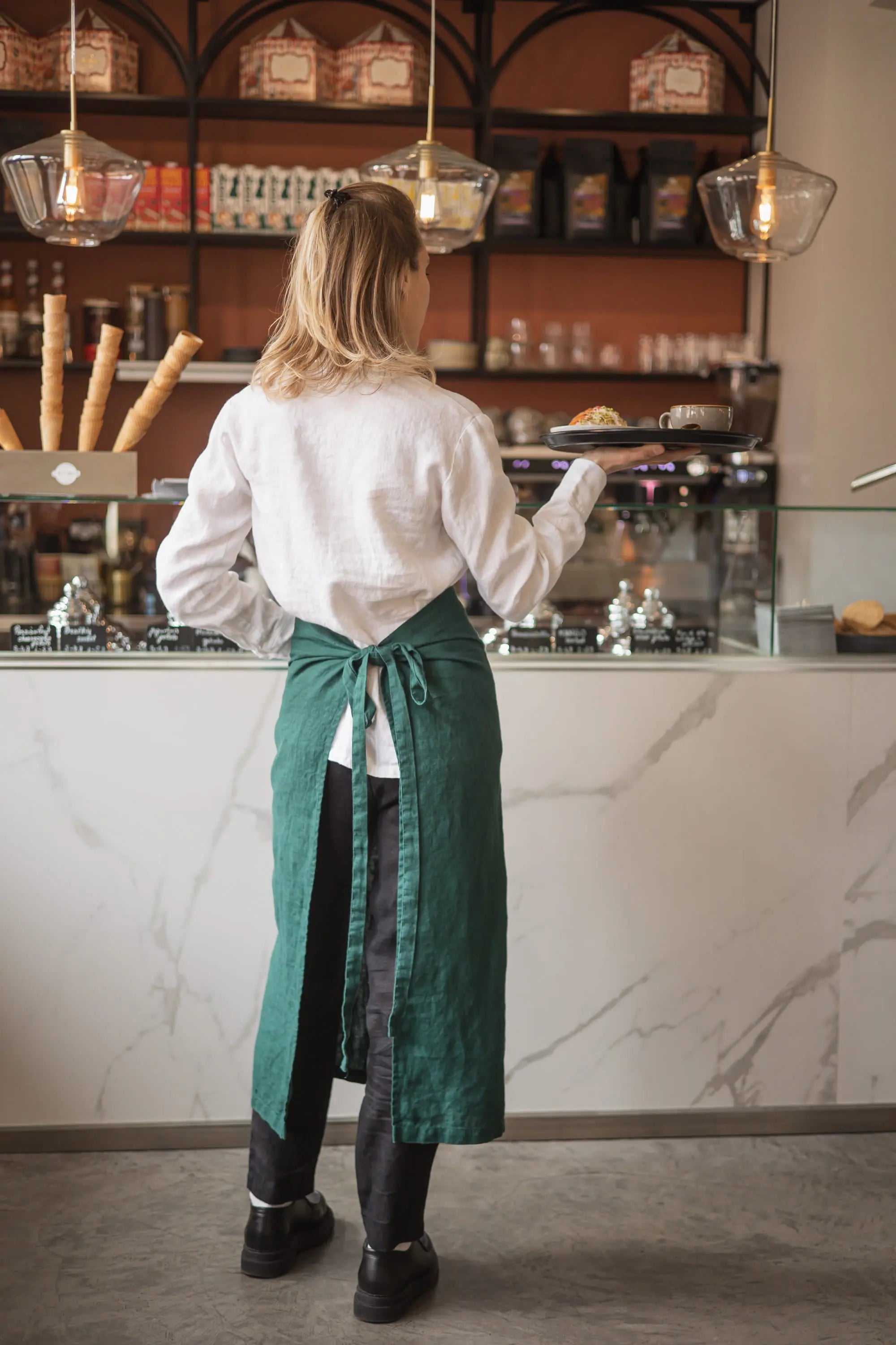 Woman viewed from behind wearing a dark green linen waist apron tied in a bow at the back, holding a serving tray while walking through a café