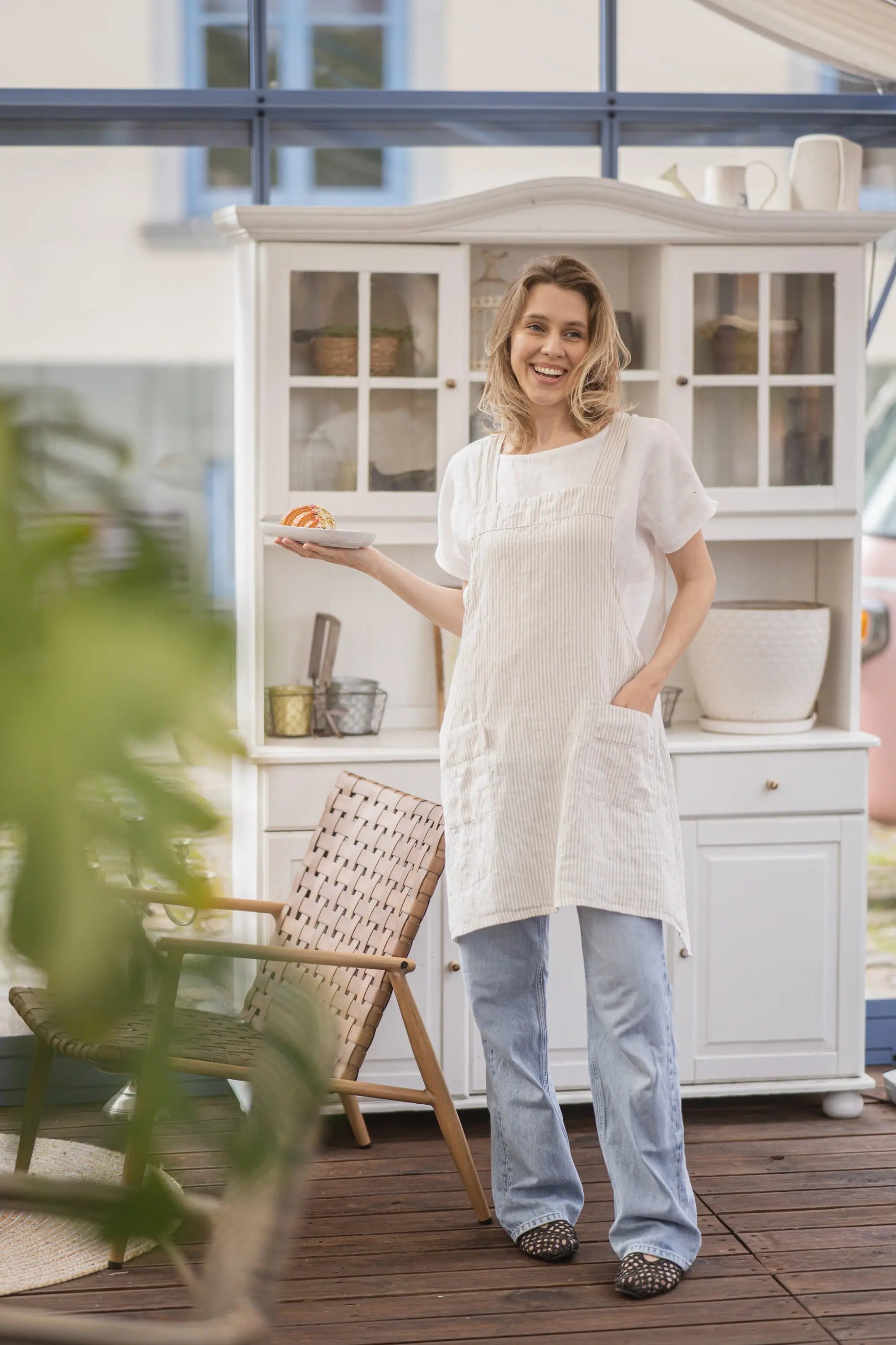 Full-length front view of a woman in a striped linen apron with front pockets, standing indoors near a white cabinet and holding a plate with a pastry.