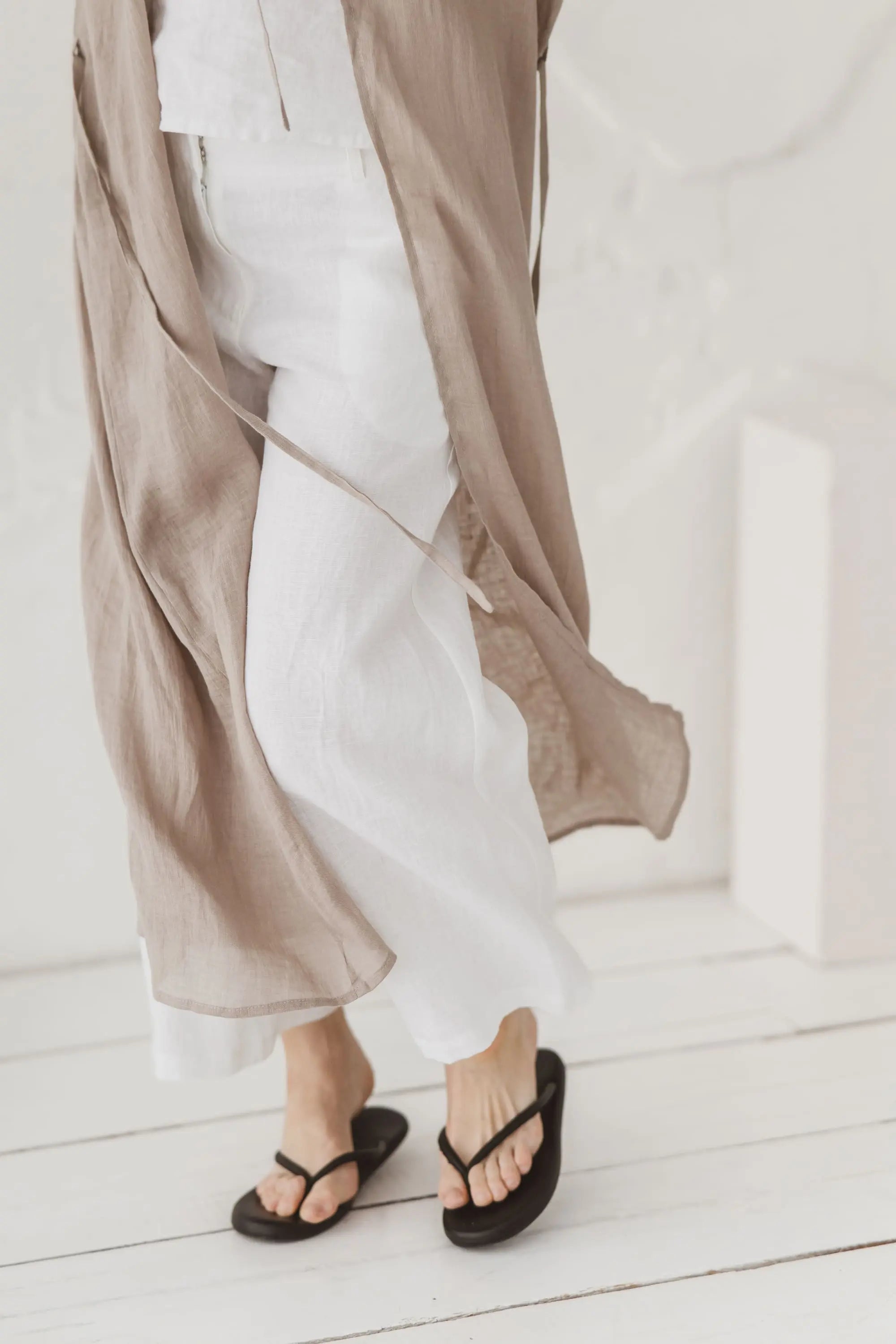 Lower half of a natural linen kaftan styled over white linen trousers with sandals