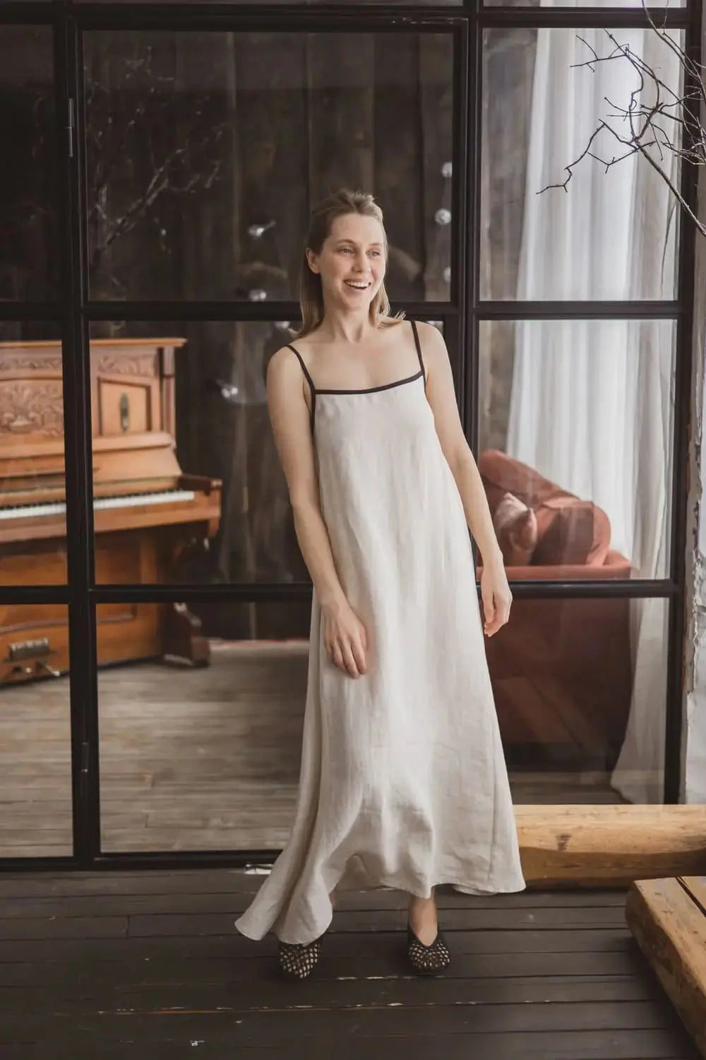 Woman smiling in a natural linen maxi slip dress with contrast black trim and spaghetti straps, full length front view