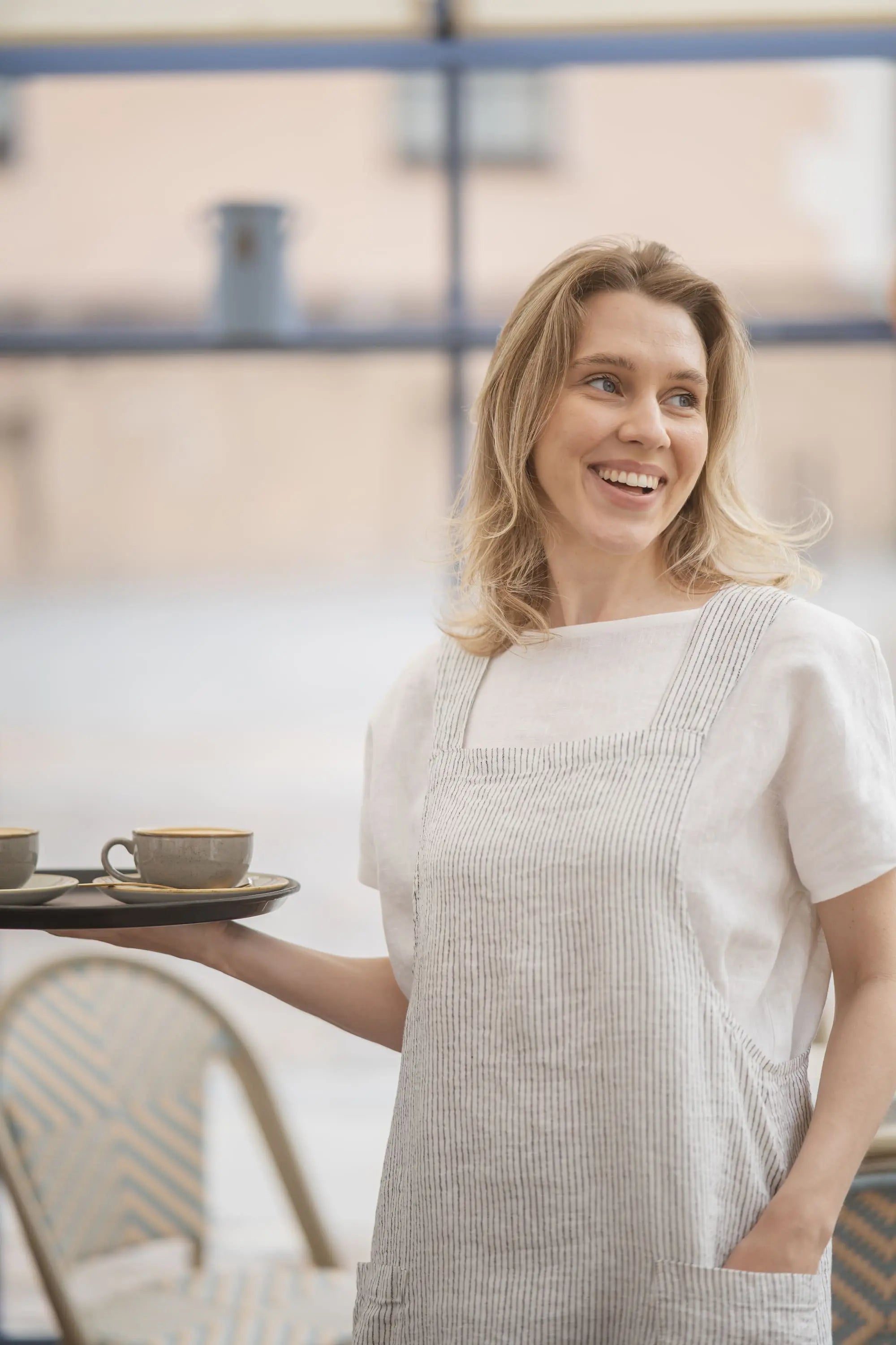 Woman in a pinstriped linen pinafore apron holding a tray with two coffee cups and saucers, smiling on an outdoor terrace.