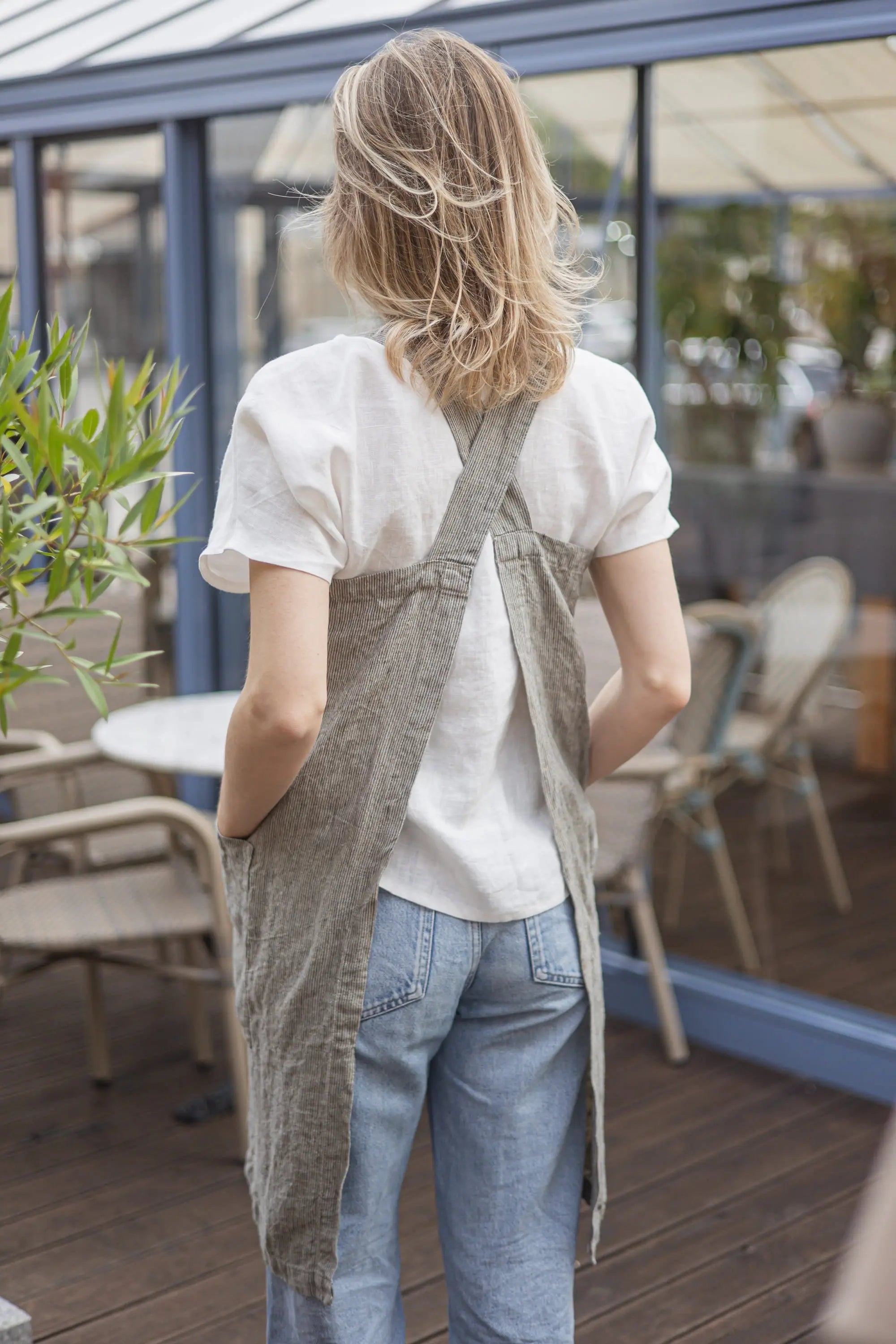 Back view close-up of a grey striped linen cross-back apron showing the crossed shoulder straps over a white t-shirt.