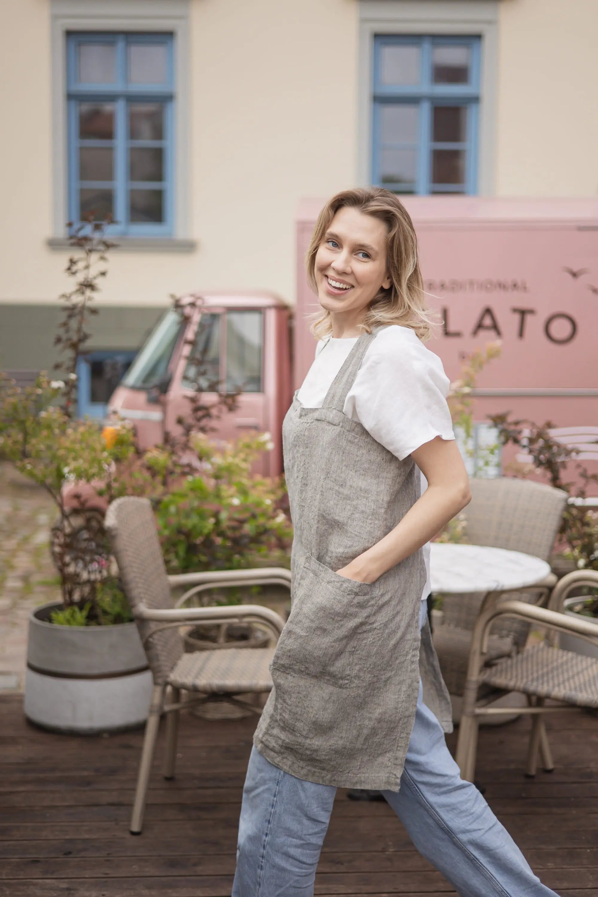 Woman walking in a grey striped linen cross-back apron, smiling with her arm raised against an open door frame outdoors.