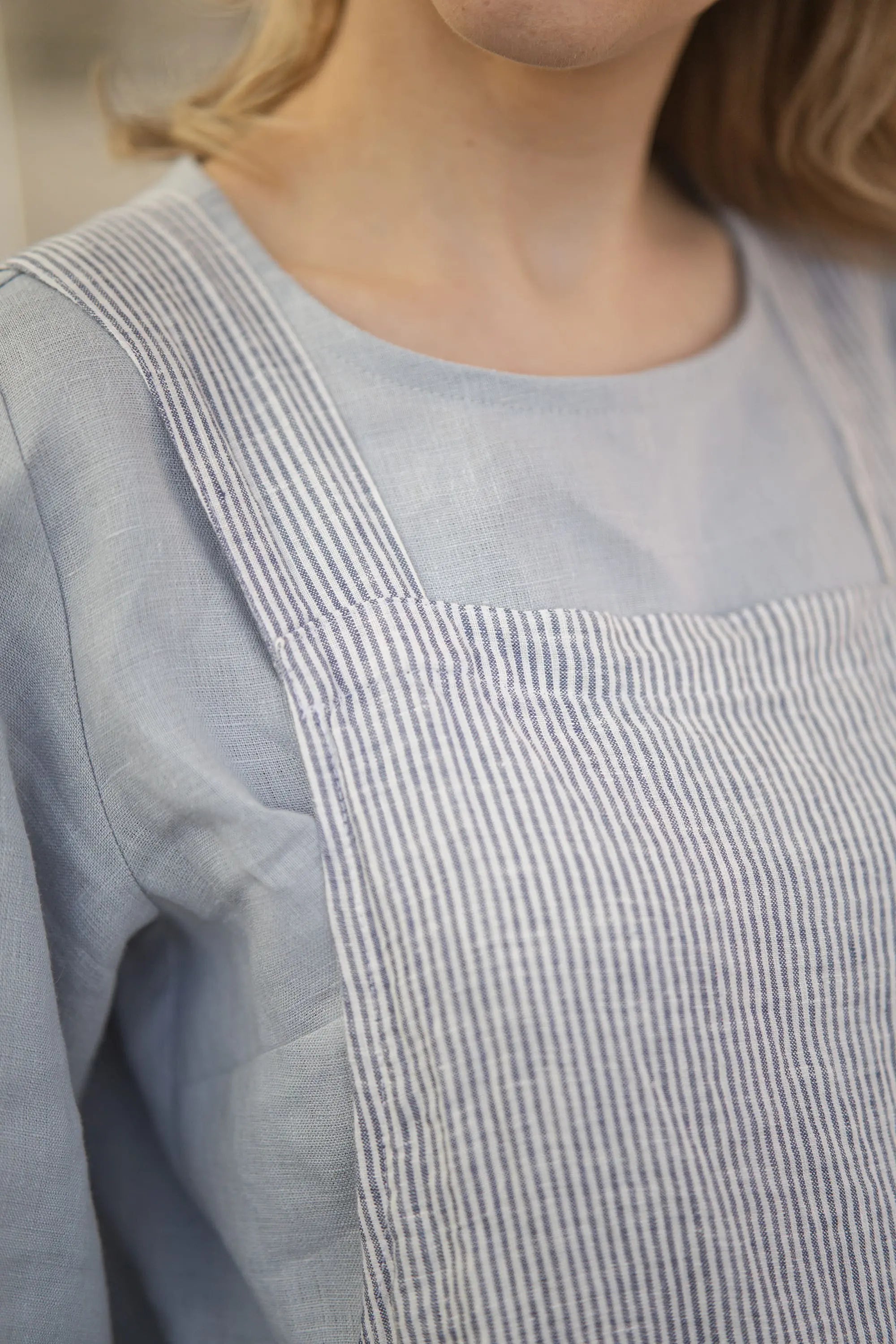 Close-up of the crossback strap and stitching detail on a striped linen apron, worn over a solid light blue-gray linen top
