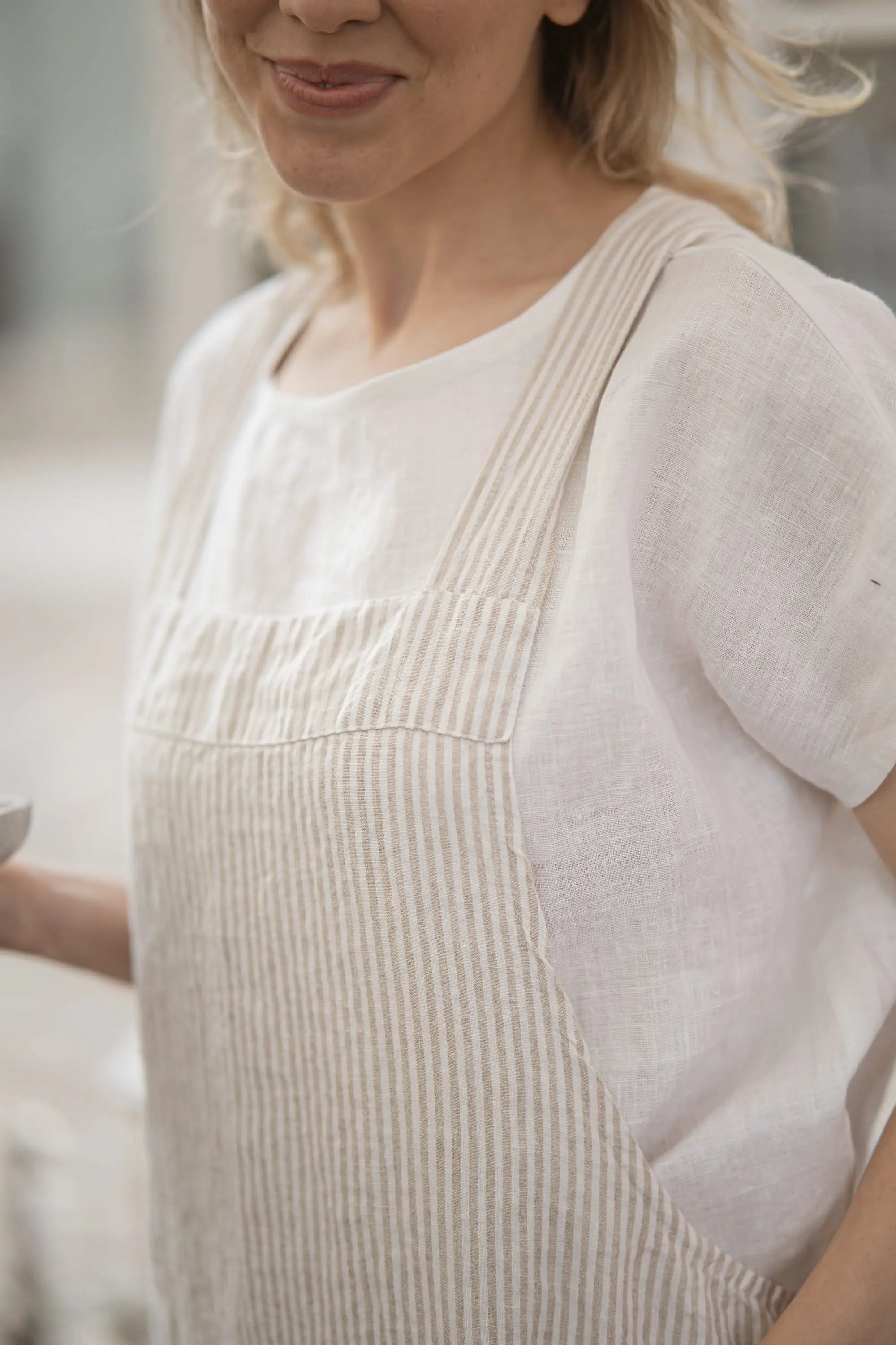 Close-up of a striped linen apron's shoulder strap and bib seam against a white top, with the woman smiling softly